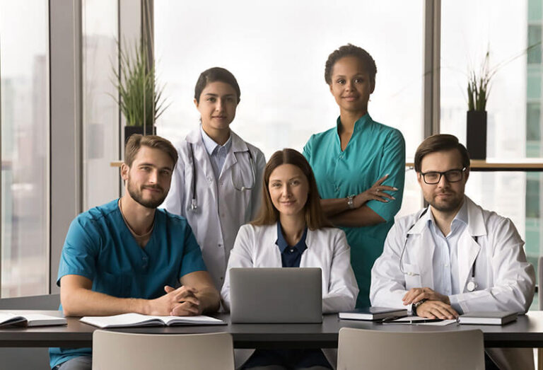 Positive diverse team of attractive young practitioners, surgeons, nurses posing together in hospital office, sitting and standing at table with laptop, looking at camera for portrait